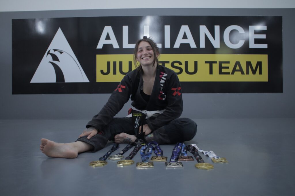 Amanda Baronio é uma mulher branca, compete na categoria meio pesado, tem cabelos castanhos escuros e tatuagens. Está sentada em um tatame com nove medalhas espalhadas no chão em sua frente. Ela sorri. Ao fundo, um painel com o texto "Alliance Jiu-Jitsu Team".