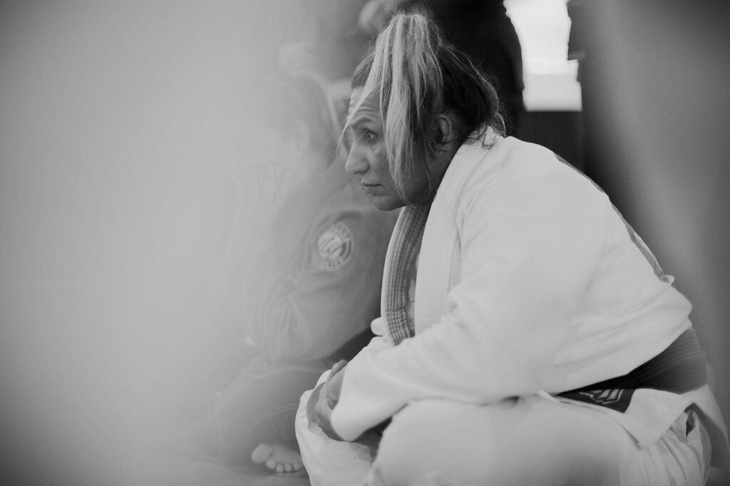 Mulher de quimono branco com cabelo preso no topo da cabeça olha atentamente para instruções de treinador.
