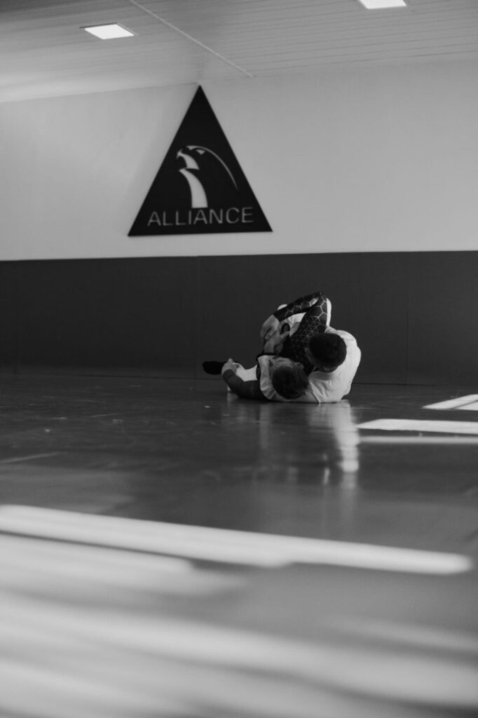 Dois atletas homens de jiu-jitsu estão em luta, deitados no tatame da Alliance Novo Hamburgo.